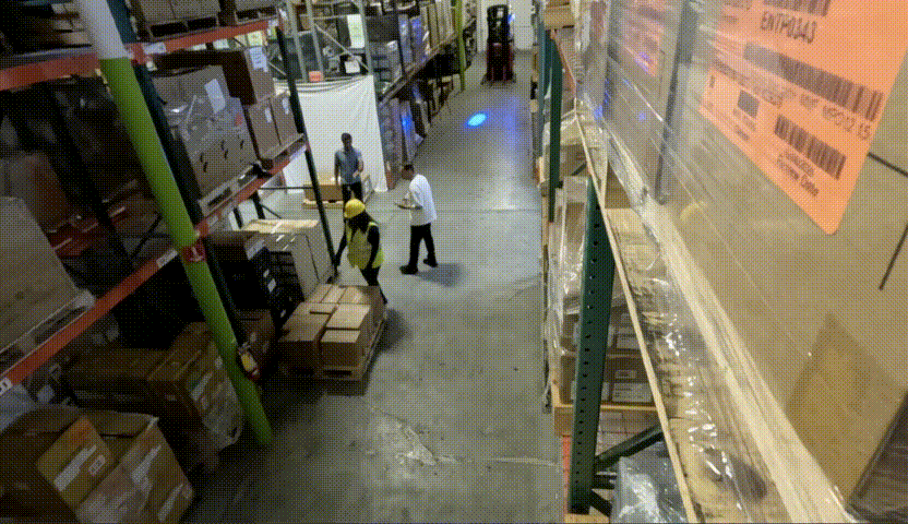 A video shows a corridor with shelves of boxes on either side and a pile of boxes on the ground. Three people are standing next to the pile of boxes. There’s a small explosion from one of the boxes on the floor, and it starts smoking. 